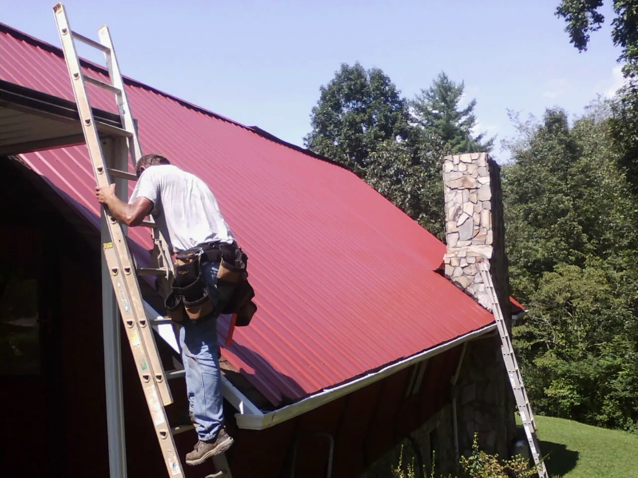Roofer on a ladder beside a red metal roof and stone chimney — Soffit & Fascia Repair by Roofing Brothers in Pleasant Plains